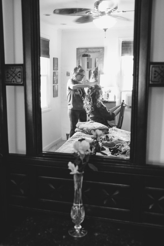 Wedding photography, a reflection shot in a mirror. A bride sits in a chair having her long curly hair styled. 