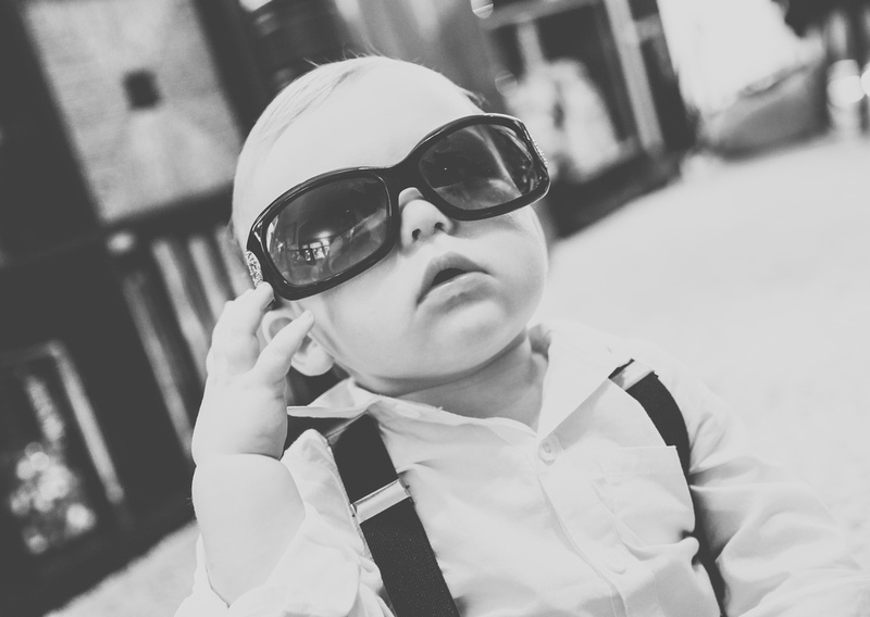 Wedding photography, a baby boy in a white shirt with suspenders is wearing oversized black sunglasses. 