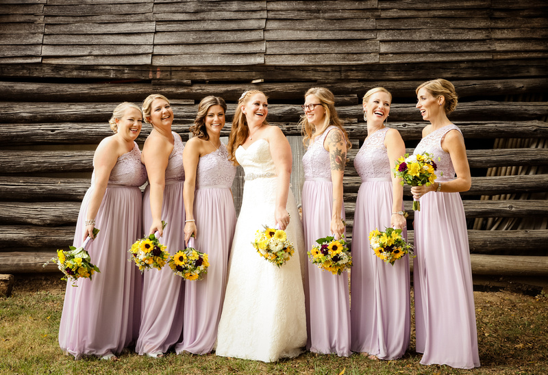 Wedding photography, a bride laughs with her bridesmaids. They are in lavender dresses and standing in front of a barn. 