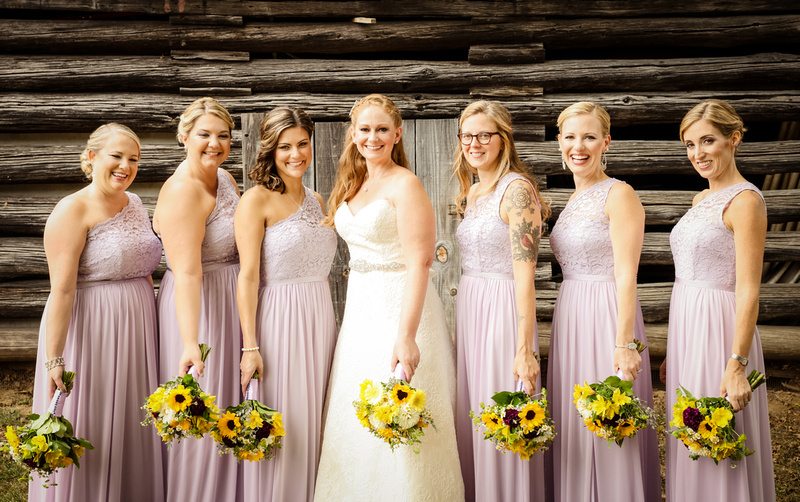 Wedding photography, a bride and her bridesmaids stand in front of a wooden barn. They wear lavender dresses and hold sunflowers. 