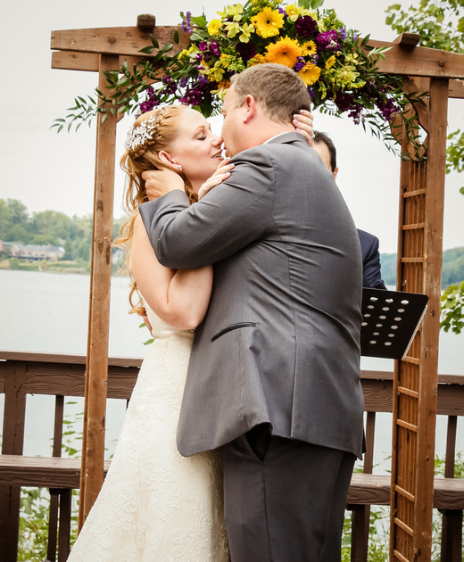 Wedding photography, the bride and groom share their first kiss. The wooden arbor behind them has purple and yellow flowers. 