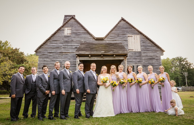 Wedding photography, the wedding party stands in front of a historic brown barn. The bridesmaids wear lavender. 