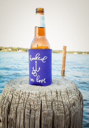 Wedding photography, a beer bottle sits on a dock in a blue koozie that says, " hooked on love,"