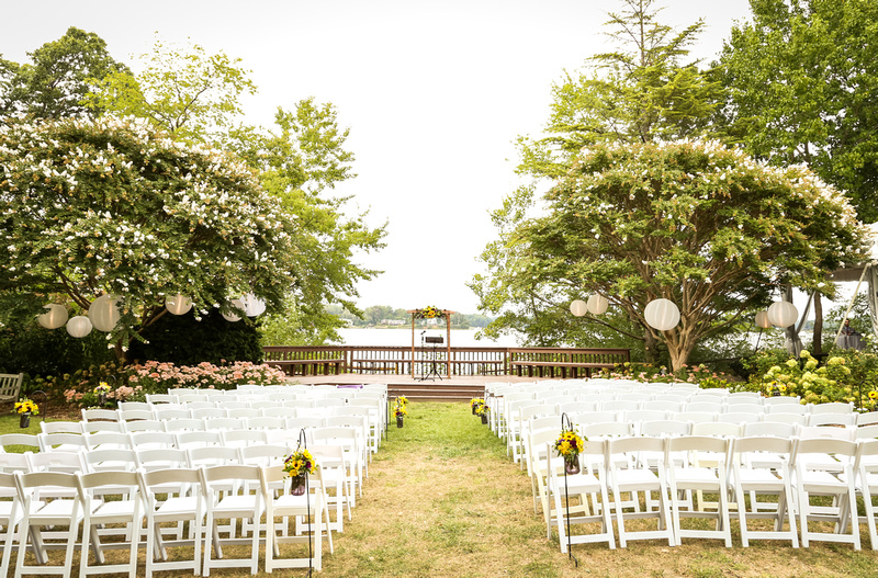 Wedding photography, a waterfront ceremony site is setup with white chairs and lanterns in the trees. 