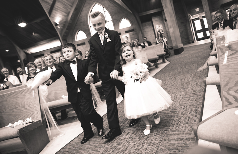 Wedding photography, two ring bearers and flower girl walk down the aisle of a church. The flower girl is sobbing for her mom. 