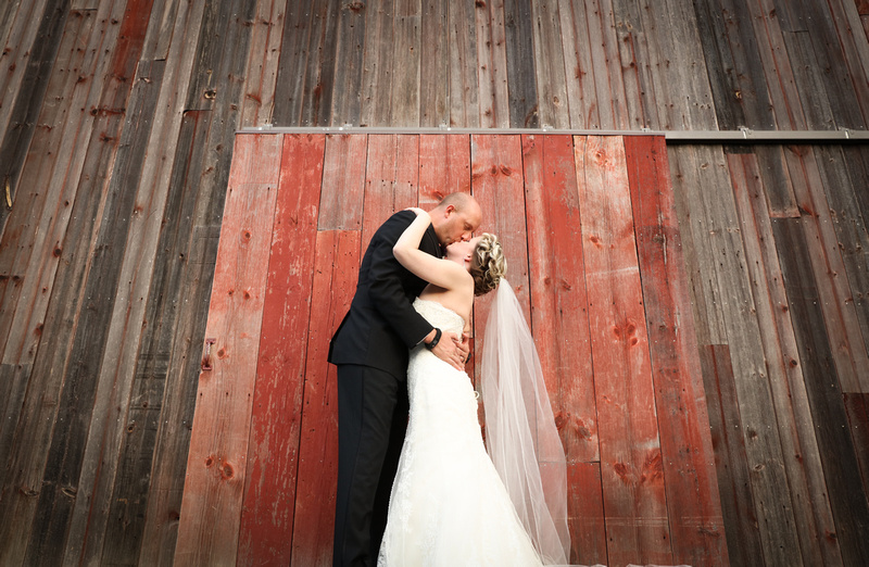 Wedding photography, the bride and groom kiss in front of brown and red aged barn doors. 