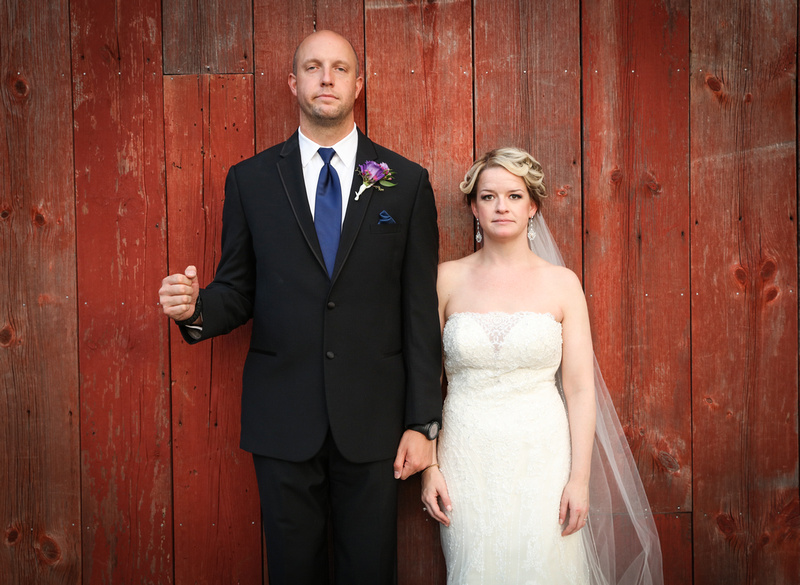 Wedding photography, the bride and groom mirror American Gothic in front of red barn doors. 