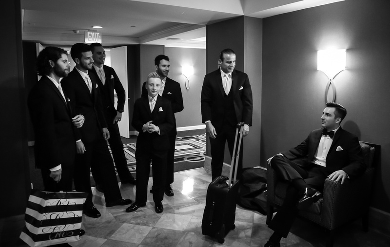 Wedding photography, a dark haired groom in a black tuxedo sits confidently in a chair while his groomsmen stand nearby smiling. 