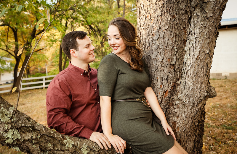 Engagement photography: a young woman in a green dress sits on a tree. Her fiancé smiles at her. 