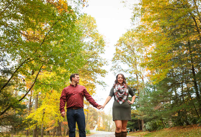 Engagement photography: fall foliage surrounds a young couple holding hands. She wears a green dress and plaid scarf. 