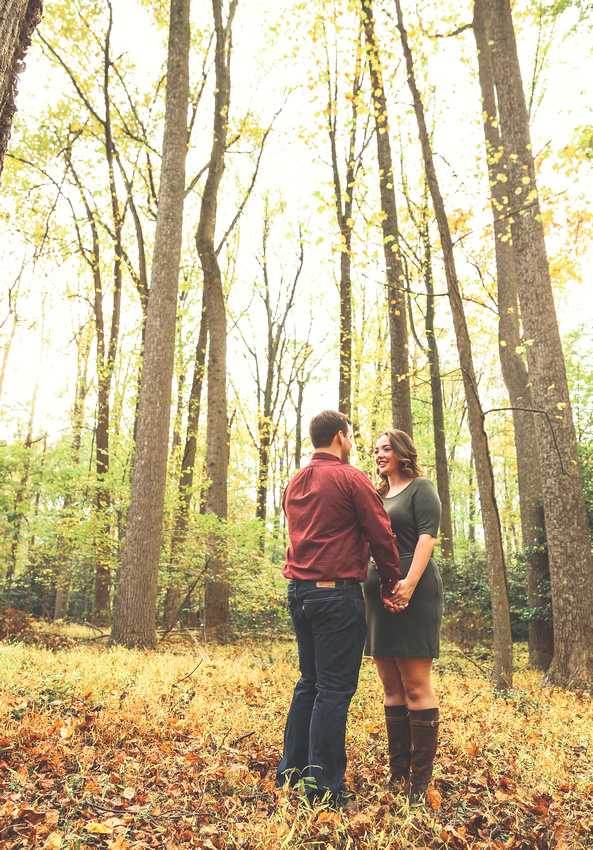Engagement photography: a young couple in the woods holds hands next to tall trees. 
