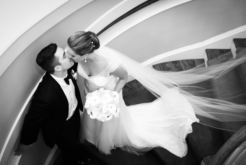 Wedding photography, a bride and groom kiss sweetly on a cascading stairwell. Her veil is draped across the stairs. 