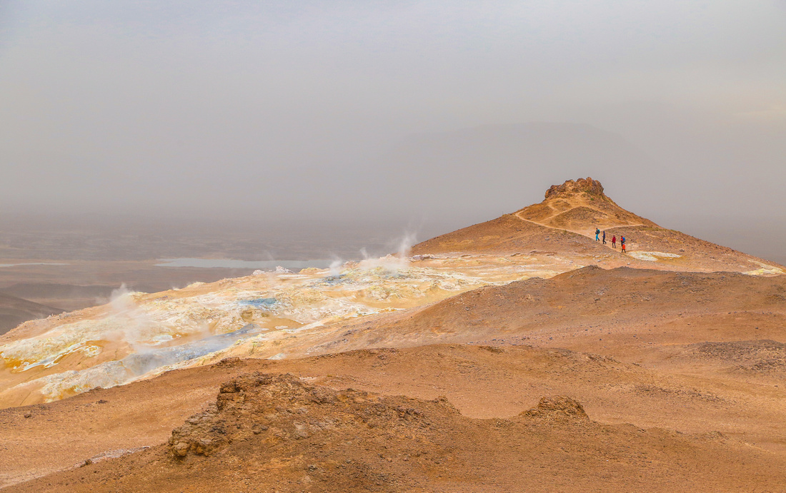 Geothermal steam rises out of the cracks of an orange-brown mountain of iceland. Far in the distance, four hikers walk the trail. 2016 in photos.
