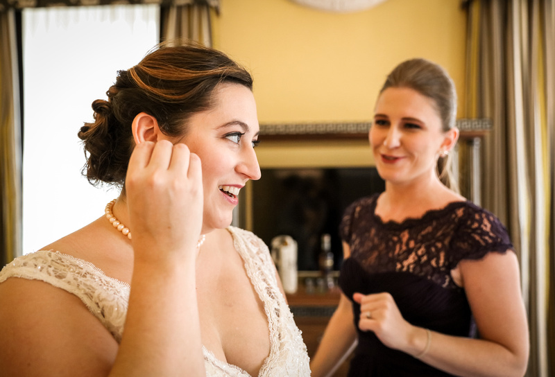 Wedding photography, a bride in a lace gown smiles while putting on her earrings. 