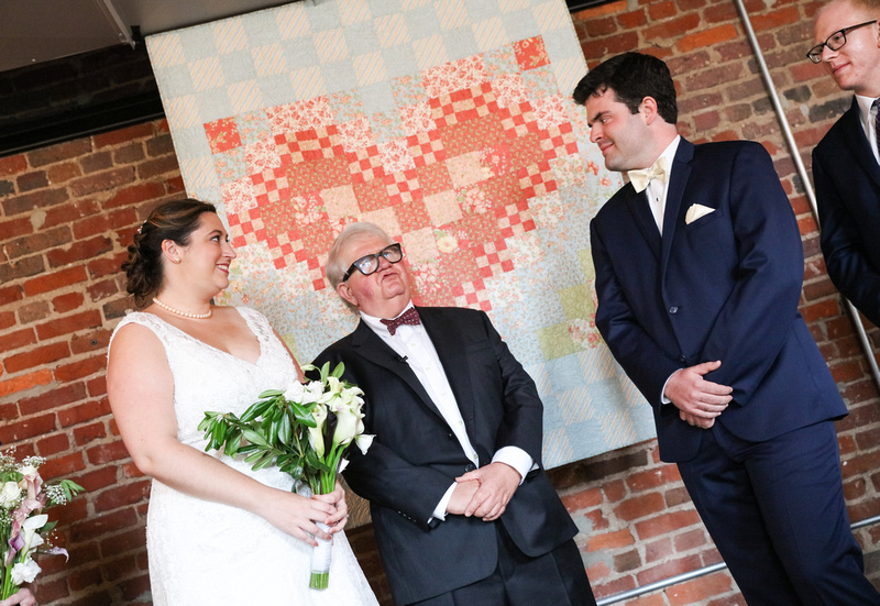 Wedding photography, a bride and groom smile during their wedding ceremony in front of a brick wall. 