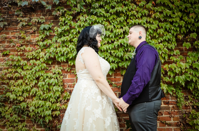 Wedding photography, a bride and groom hold hands lovingly in front of a brick wall with green ivy. 