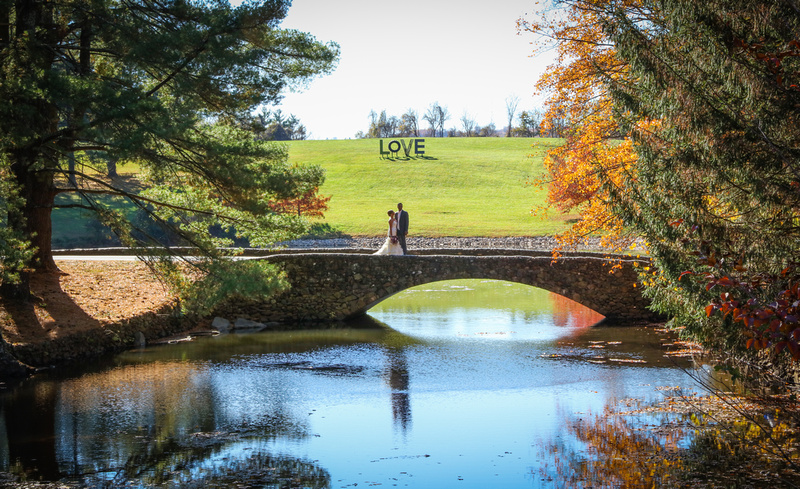 Wedding photography, a bride and groom stand on a stone bridge. A sign reading love is on the hill behind them. 