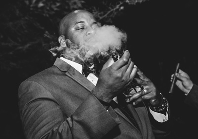 Wedding photography, a black and white image of a groomsman lighting a cigar with a big puff of smoke. 