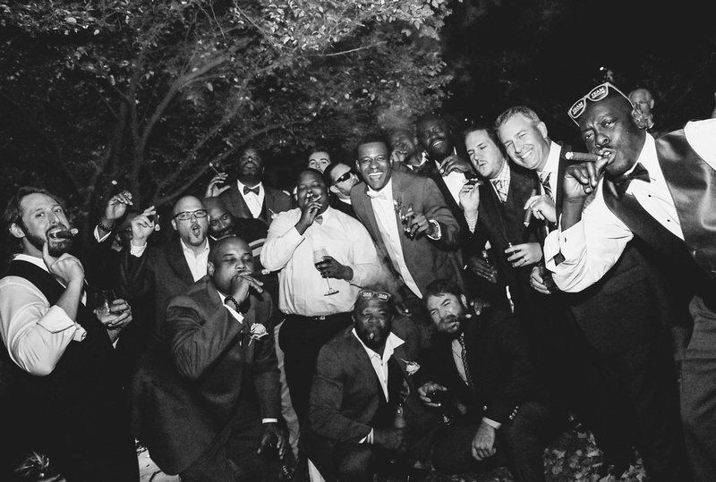 Wedding photography, a black and white image of a big group laughing while smoking cigars under a tree. 