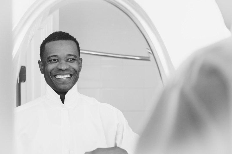 Wedding photography, a black and white image of a groom smiling in the mirror as he buttons his shirt. 
