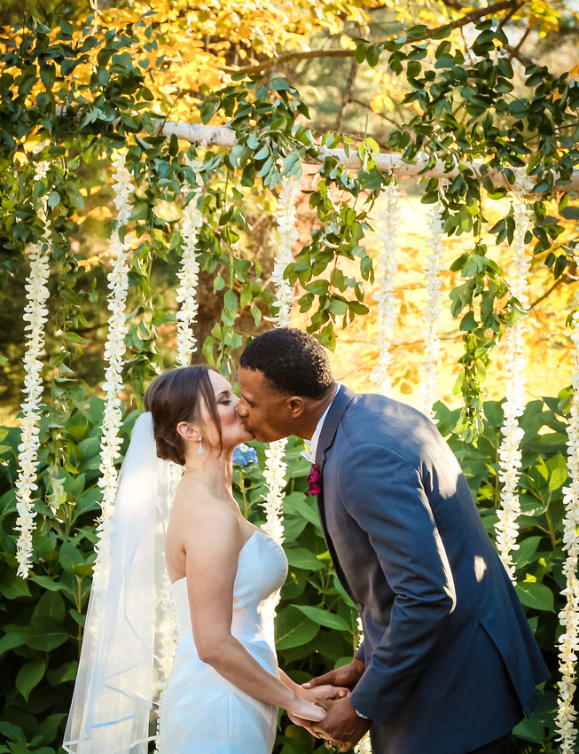 Wedding photography, the bride and groom share their first kiss as husband and wife. Flower petals hang behind them. 