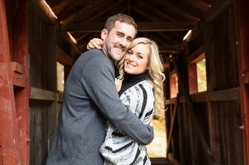 Engagement photography, a blond woman and man embrace under a red covered bridge. 