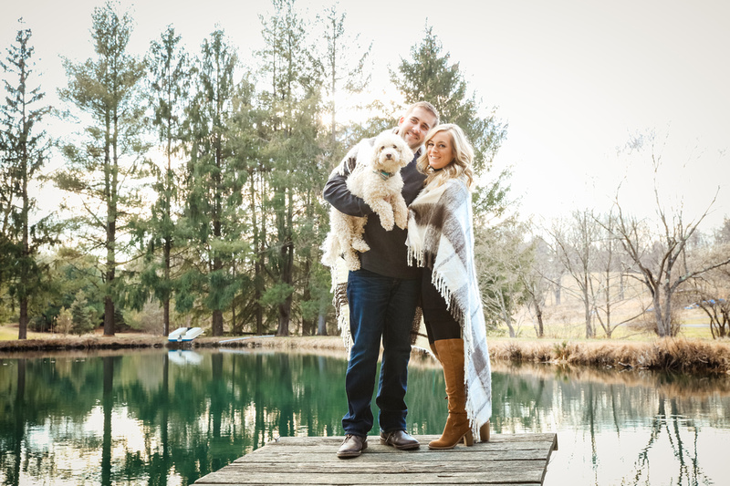 Engagement photography, a couple stands on a dock in front of a pond holding a small white dog. 