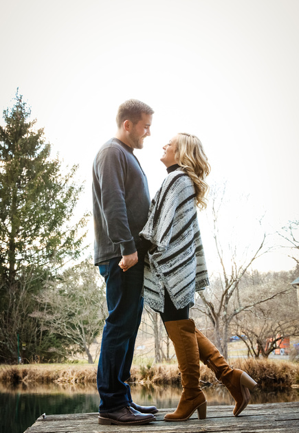 Engagement photography, a blond woman in brown knee-high boots smiles at her fiancé. They stand on a dock. 