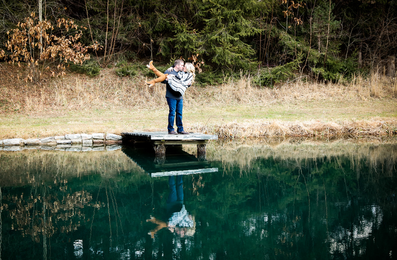 Engagement photography, a man picks up his fiancé on a dock and kisses her. They are in the woods. 