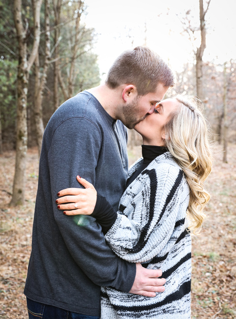 Engagement photography, a couple kisses passionately in the woods. She wear a black and grey sweater. 