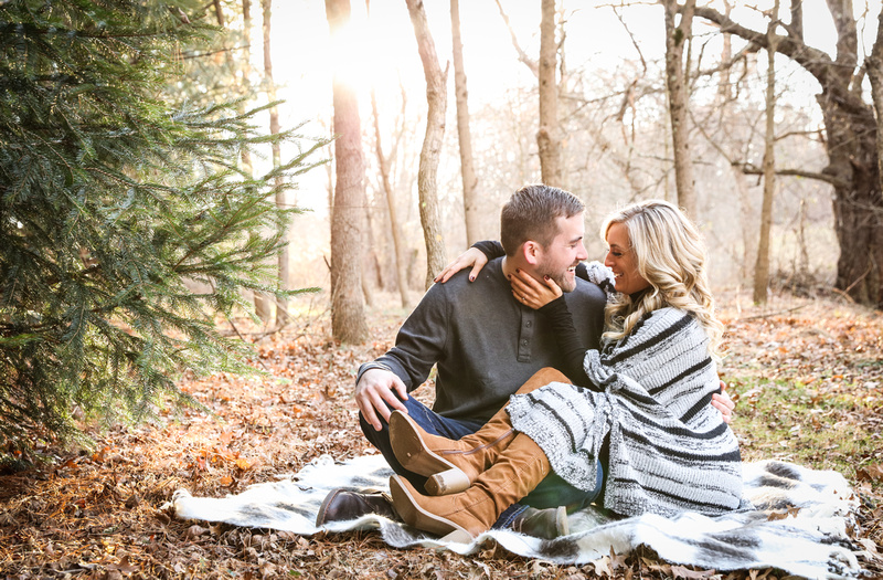 Engagement photography, a couple sits on a blanket in the woods on a sunny winter day. They are laughing. 