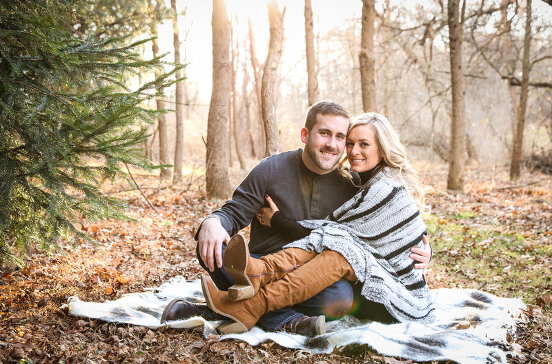 Engagement photography, a couple smiles at the camera while sitting on a blanket in the woods. 