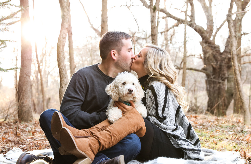 Engagement photography, a couple sits in the woods kissing with their small white dog on their laps. 