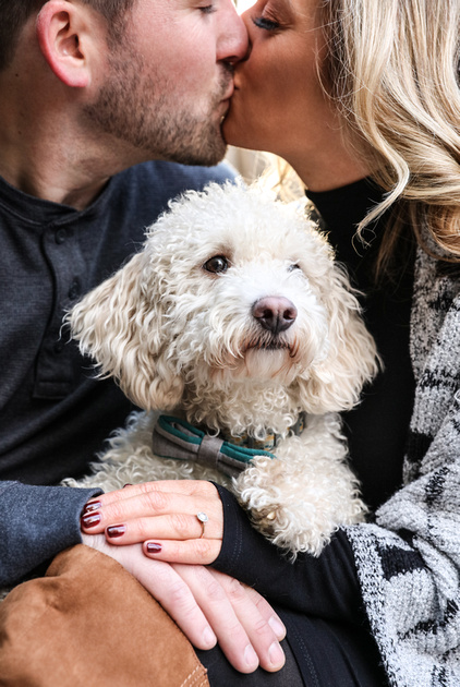 Engagement photography, a small white dog sits with his owners while they kiss. 