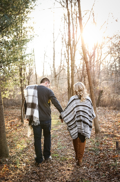 Engagement photography, a couple walks in the woods holding hands. The man has a blanket over his shoulder. 