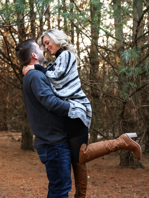 Engagement photography, a man picks up his fiancé and they smile at each other. She wears brown knee-high boots. 