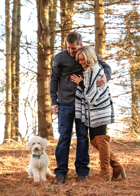 Engagement photography, a couple stands together in the woods looking down at their small white dog. 