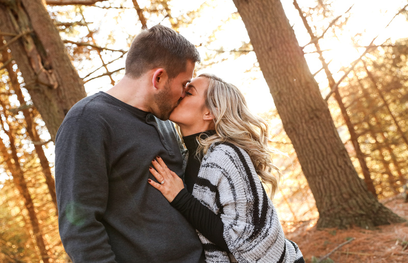 Engagement photography, a couple kisses sweetly in the woods with sunshine pouring through the trees. 