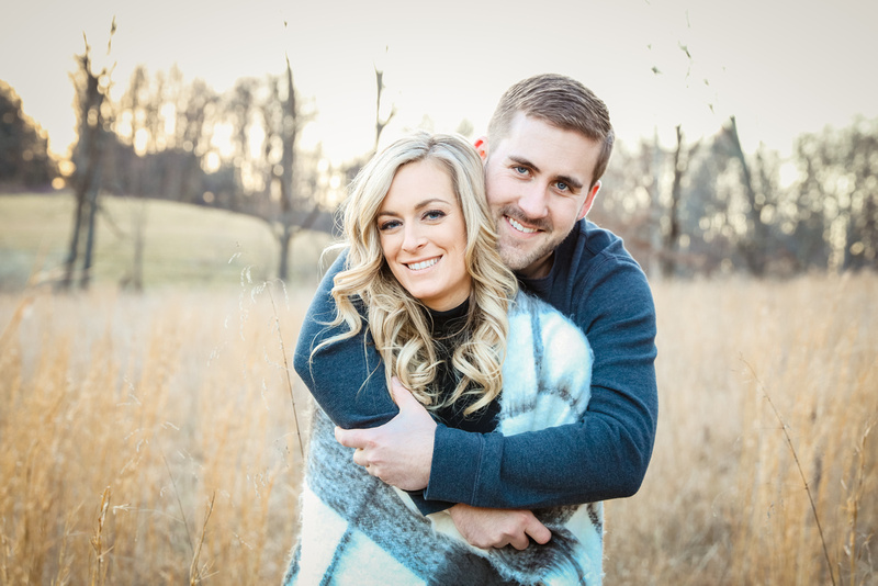 Engagement photography, a man wraps up her fiancé in a grey blanket. They stand in a field. 