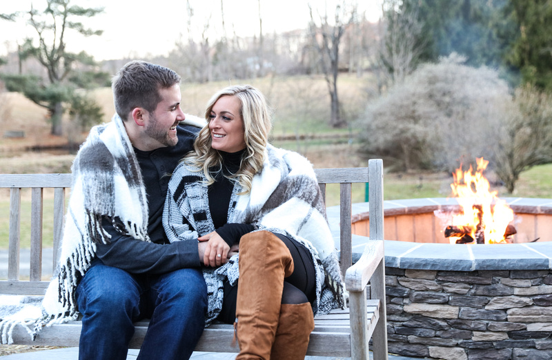 Engagement photography, a couple snuggles on a bench, sitting by an outdoor fire pit. 