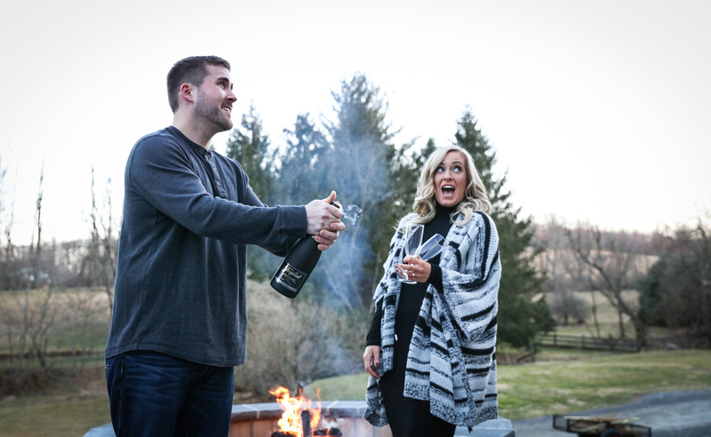 Engagement photography, a couple is surprised as they pop open a bottle of champagne by a fire pit. 