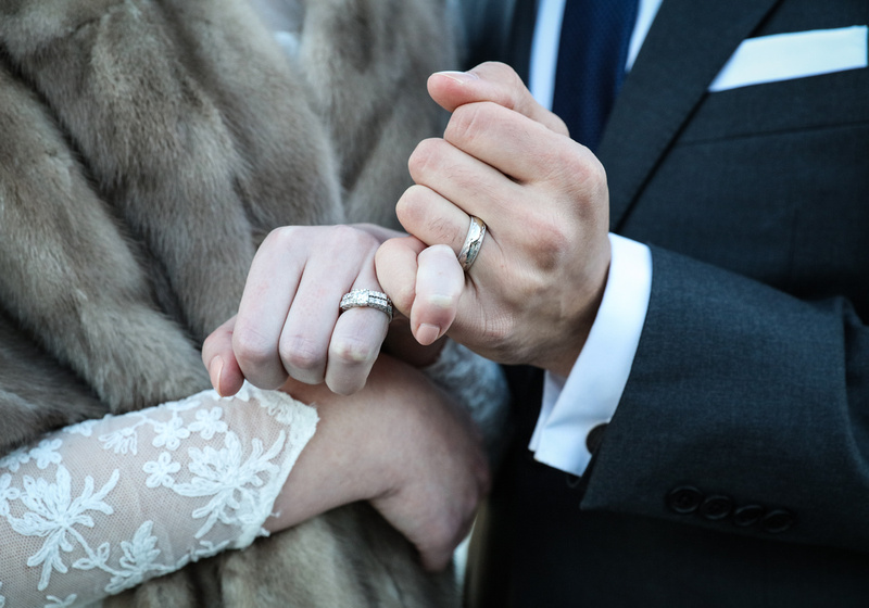 Wedding photography, a closeup of a bride and groom linking fingers with their wedding bands. He wears a grey suit, she wear lace and fur. 