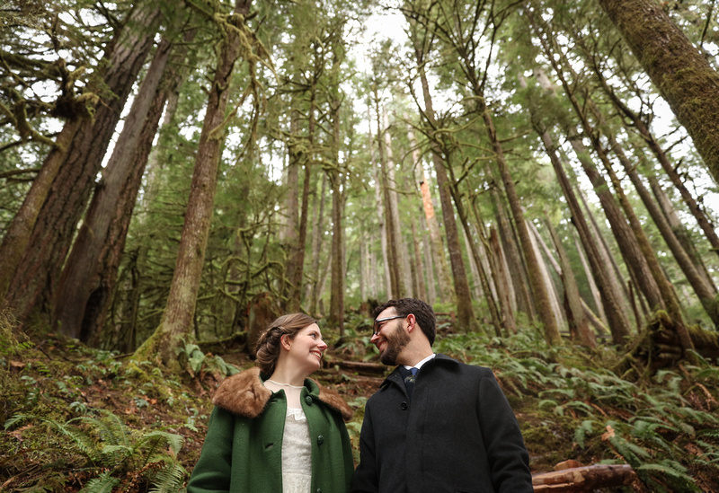 Wedding photography: a bride in a green coat and groom look at each other and smile. They are surrounded by a forest of trees. 