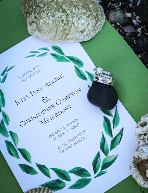 Wedding photography, a wedding invitation with green accents and seashells. The wedding rings balance on a rock. 