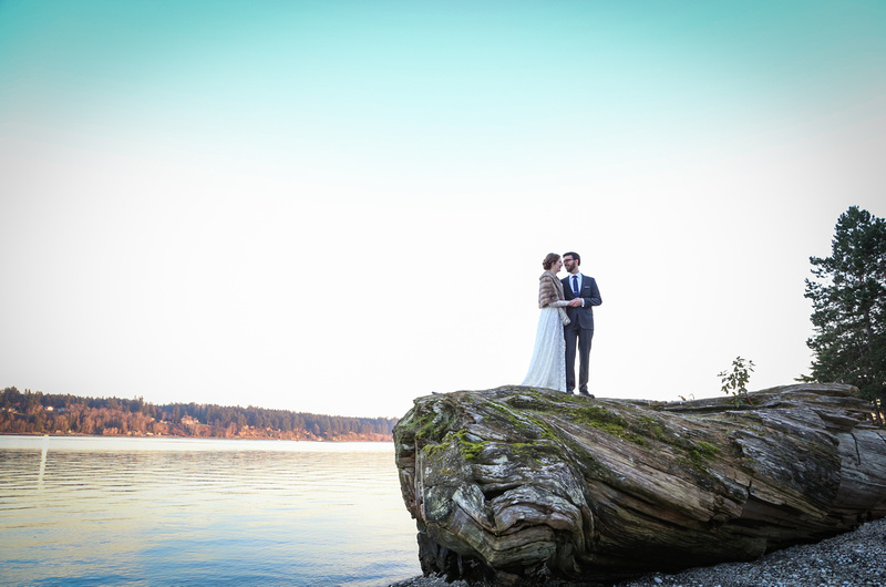 Wedding photography, a bride and groom stand on a giant piece of driftwood by the water, at sunset. 