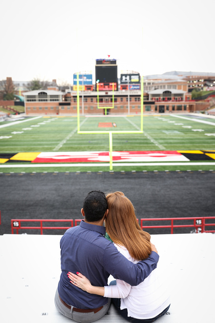 Engagement photography, a couple sits in the bleachers with their arms around each other. He wears a blue shirt. 