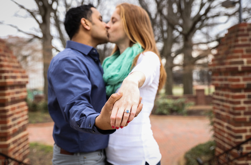 Engagement photography, a couple kisses while holding out their hands. She wears a ring with a blue stone. 