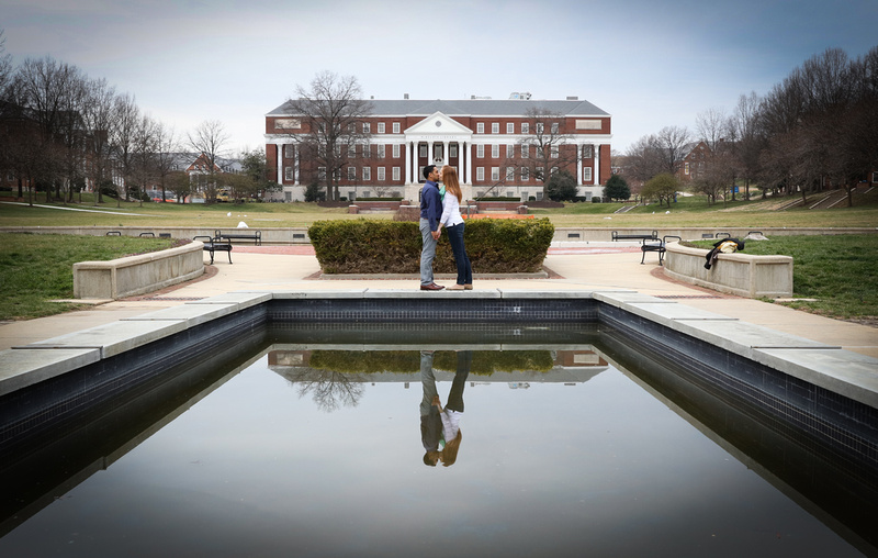 Engagement photography, a couple kisses in front of a reflective pool of water on a school campus. 