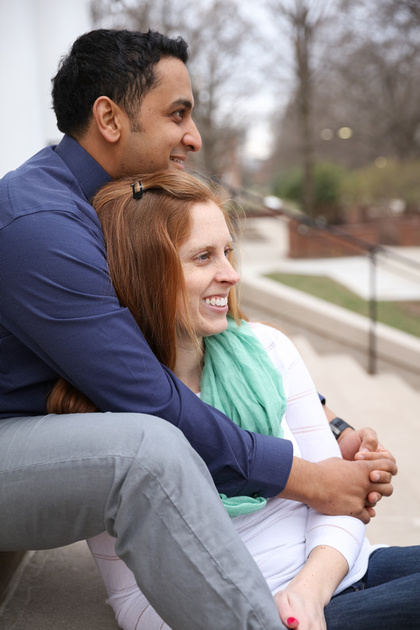 Engagement photography, a woman sits on the steps between her fiancé's legs. She has red hair, he is Indian. 
