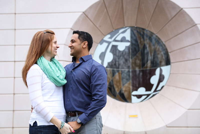 Engagement photography, a couple smiles at each other in front of a window with the Maryland flag symbol on it. 
